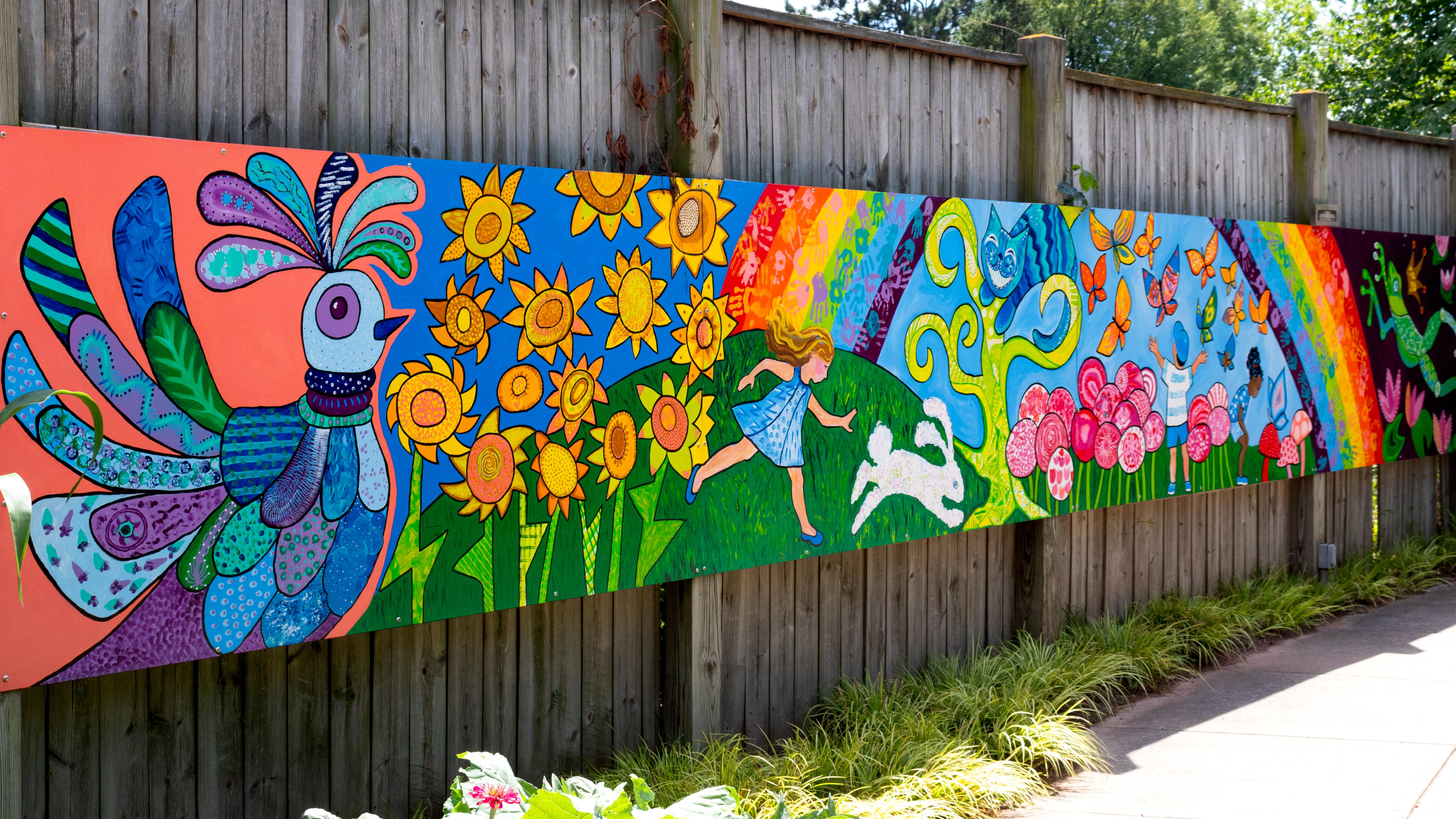 Colorful mural with a bird, sunflowers, a girl, a rabbit, a rainbow, a cat, butterflies, and flowers on a wooden fence